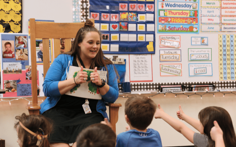 A kindergarten teacher is sitting in a chair holding a book and giving two thumbs up to her students.